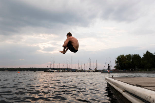 Rear view of a young girl mid-air as she has just run and jumped off the end of the dock at a lake. Taken on a beautiful summer day. The girl is wearing a pink life jacket with her pink and purple swim wear and black water shoes.