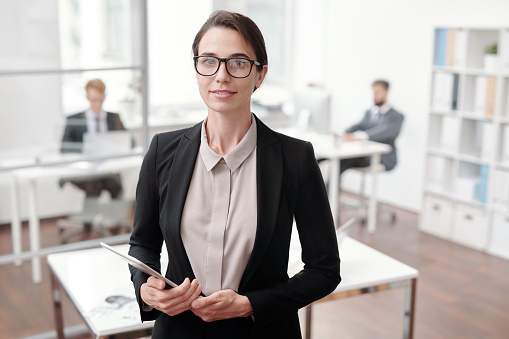 Waist up portrait of young businesswoman looking at camera and holding digital tablet while standing in office, copy space