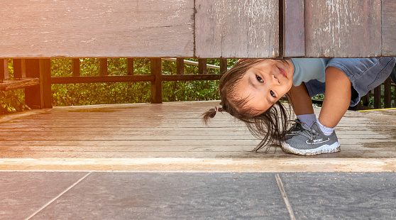 Happy Asian little child girl plays peekaboo under the wooden door in the garden.