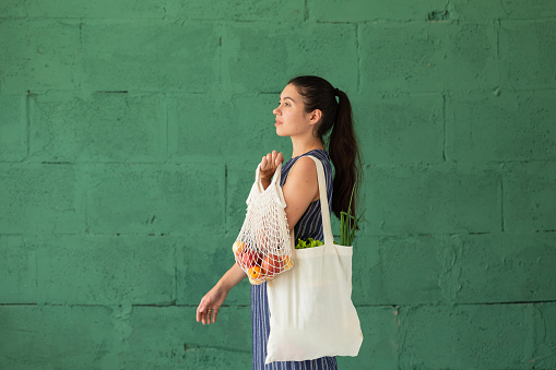 Young woman with shopping cotton Eco bag with fruits and vegetables in her hands on green wall background. Lifestyle, zero waste concept