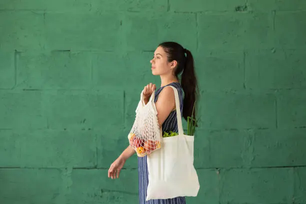 Photo of Young woman with shopping cotton Eco bag with fruits and vegetables in her hands on green wall background. Lifestyle, zero waste concept