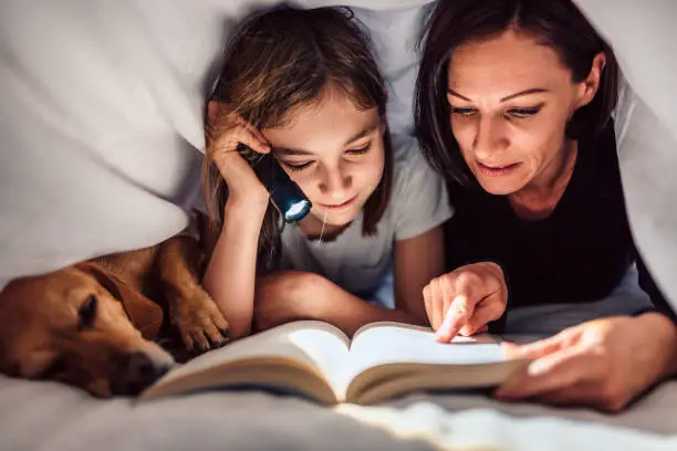Mother, daughter and dog laying on the bed under blanket holding flashlight and reading book late at night