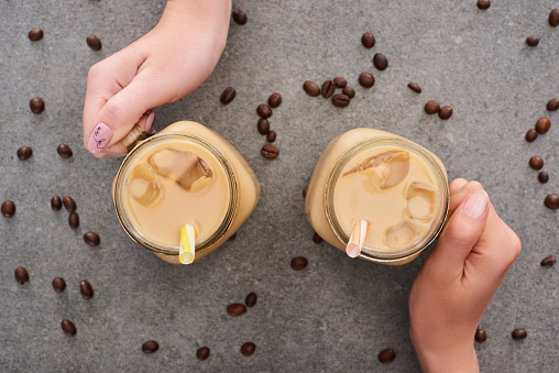 cropped view of women holding glass jars with ice coffee and coffee grains on grey background
