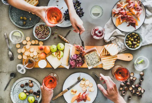 Mid-summer picnic with wine and snacks. Flat-lay of charcuterie and cheese board, rose wine, nuts, olives and peoples hands holding wineglasses and celebrating over concrete table background, top view