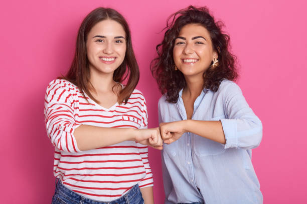 Smiling caucasian women fist bumping in studio, two friends greeting or supporting team motivation, females wear casual shirtts, have darl hair, looks at camera with toothy smile, expresse happyness. sonriendo mujeres caucásicas puño golpeo en el estudio, dos amigos saludando o apoyando la motivación del equipo, las mujeres usan camisetas casuales, tienen el pelo atrevido, mira la cámara con la sonrisa dentada, expresan la felicidad.