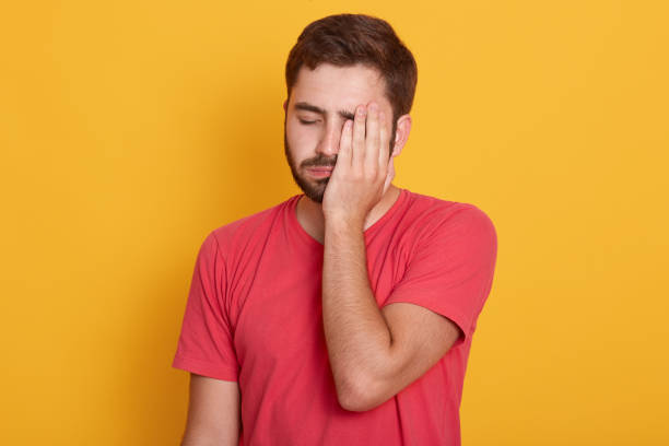 istock Exhausted young unshaven ing half of face with hand while standing against yellow studio background, looks tired, feeling depressed, has problems, posing with closed eyes, dressed red t shirt. 1170483251