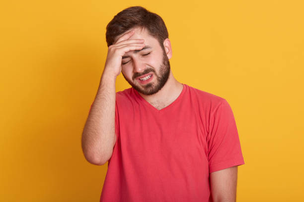 istock Tired young man keeps hand on forehead, handsome guy wearing red casual t shirt, posing with closed eyes over yellow studio background, suffers from headache. Stress or problem concept. Front view. 1170483255