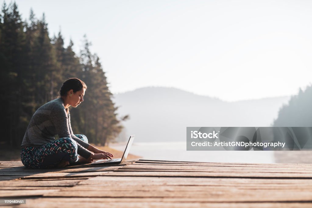 Woman using laptop outdoor at wooden pier. Woman using laptop in nature, she sitting on wooden pier around beautiful mountain lake and using laptop. Remote Location Stock Photo