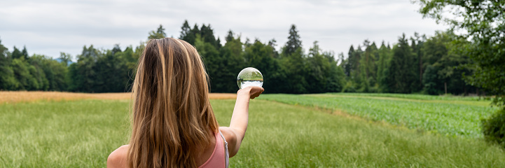 View from behind of a woman standing in nature surrounded by fields and meadows holding crystal ball in her hand.