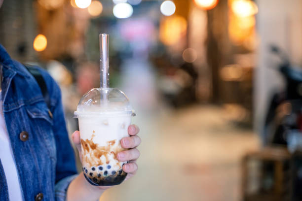 istock Young girl in denim jacket is drinking brown sugar flavored tapioca pearl bubble milk tea with glass straw in night market of Taiwan. 1170489848