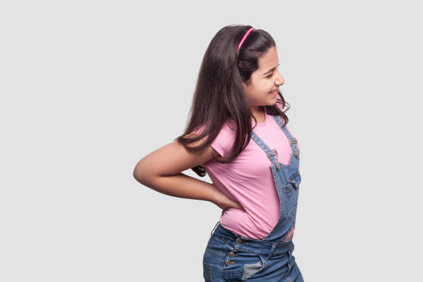 ryggrad ryggvärk. profilsida visa porträtt av sjukt brunett ung flicka i rosa t-shirt och blå overaller stående och hålla henne smärtsam rygg. - iranian girl bildbanksfoton och bilder