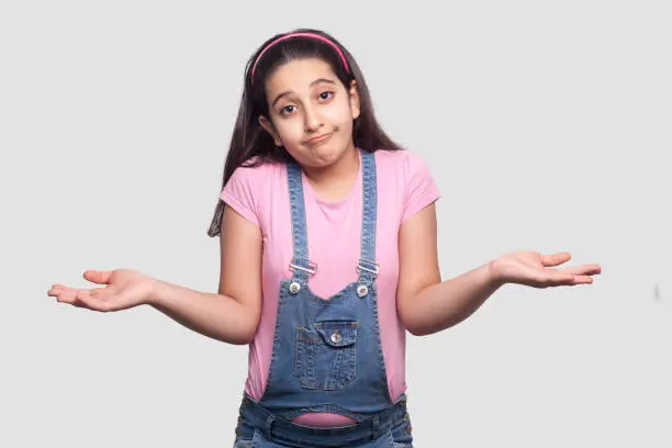 I don't know. Portrait of confused brunette young girl in casual style, pink t-shirt and blue overalls standing, raised arms and looking at camera. indoor studio shot isolated on light gray background