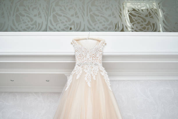 beautiful beige embroidered wedding dress hanging on hanger against wall in luxury hotel room, copy space, close up. bridal morning preparations. wedding concept - love hanging indoors studio shot imagens e fotografias de stock