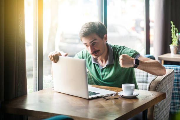 homem de negócios irritado novo no t-shirt verde que senta-se com face agressiva e punhos do encaixotamento e tentativa a perfurar seu portátil. - boxing caucasian men business - fotografias e filmes do acervo