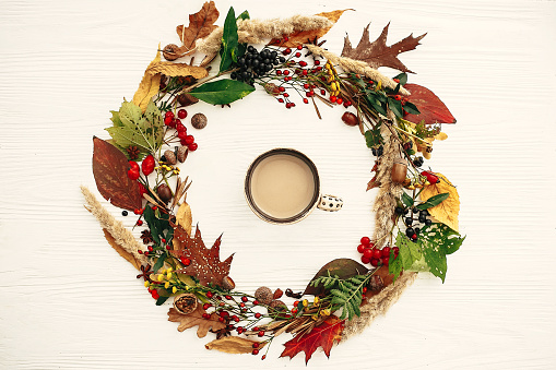 Autumn flat lay. Coffee cup in autumn wreath of fall leaves, red berries, acorns, anise, nuts, autumn flowers on white wood. Copy space.  Cozy autumn mood. Seasons greeting card
