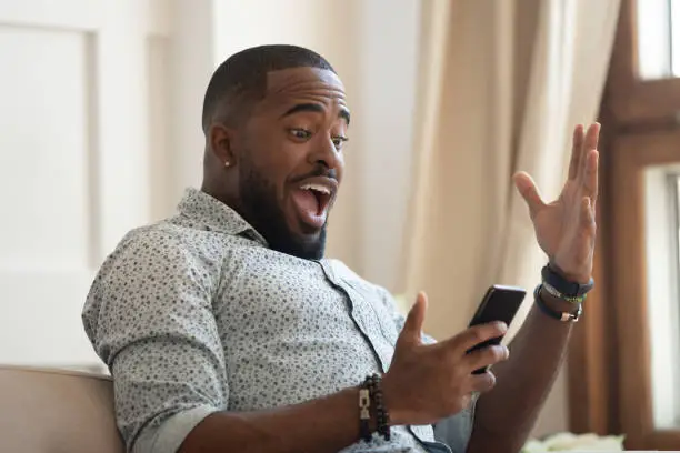 Surprised happy african man holding phone looking at cellphone read good news in sms sit on sofa, amazed black guy winner excited by mobile app win scream with joy celebrate victory success at home