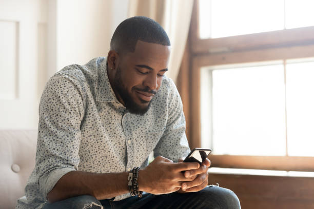 joven afroamericano usando aplicación de teléfono inteligente sentarse en el sofá - internet dating fotografías e imágenes de stock