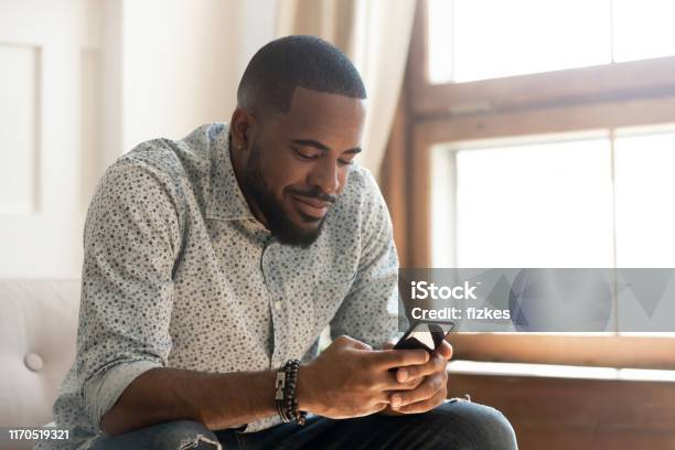 Young African American Man Using Smartphone App Sit On Sofa Stock Photo - Download Image Now