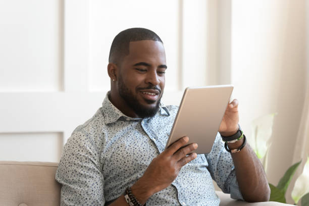 uomo africano sorridente che usa il tablet digitale che legge l'e-book a casa - reading book men sofa foto e immagini stock