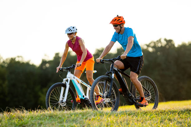 happy mature woman and man cycling together happy smiling sporty fit couple enjoying summer cycling day outdoors in rural landscape on electric mountain bikes shallow focus on man electric bicycle photos stock pictures, royalty-free photos & images