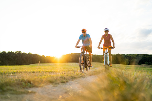 low angle view athletic sporty fit woman and man couple friends in their fourties cycling with their electric mountain bicycles on gravel path through grassland rural landscape on sunny summer afternoon sunset