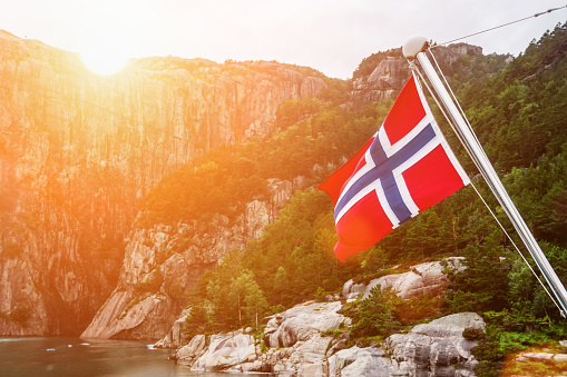 Norwegian flag on the fjord surrounded by mountains. Sunny weather. Tourism, tourist destination.