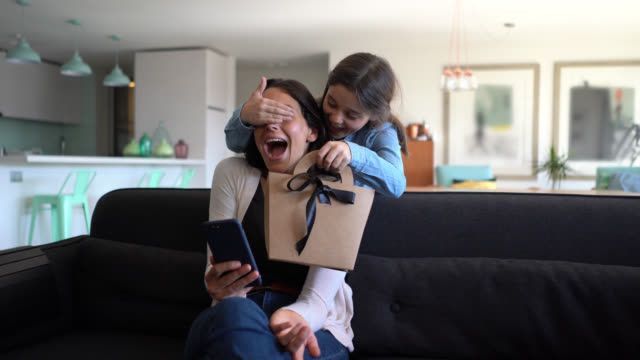 Beautiful little girl surprising her mom while she is sitting on couch using her smartphone with a gift for mother’s day