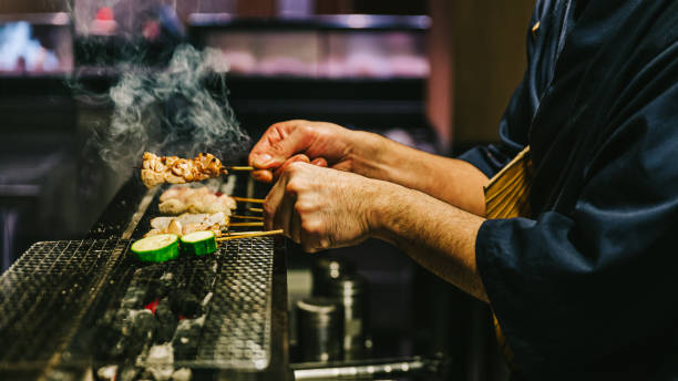 Hands of Japanese Yakitori Chef grilling chicken marinated with ginger, garlic and soy sauce and cucumber with a lot of smoke. Hands of Japanese Yakitori Chef grilling chicken marinated with ginger, garlic and soy sauce and cucumber with a lot of smoke. japanese chef stock pictures, royalty-free photos & images