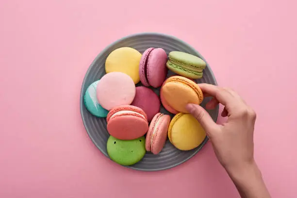 cropped view of female hand near plate with multicolored delicious French macaroons on pink background