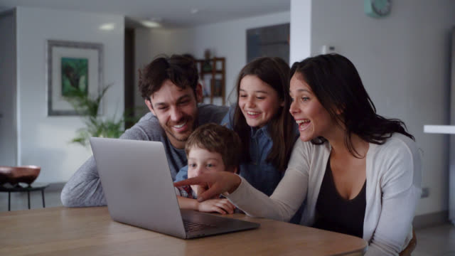 Happy family looking at videos on laptop while kids point at screen and talk smiling - Lifestyles