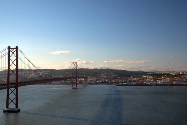 Horizontal photo Bridge April 25-de Abril, connecting Lisbon on North and Almada on the South Bank of Tagus river. Sailing boat in the Tagus River with the Lisbon skyline on the backround