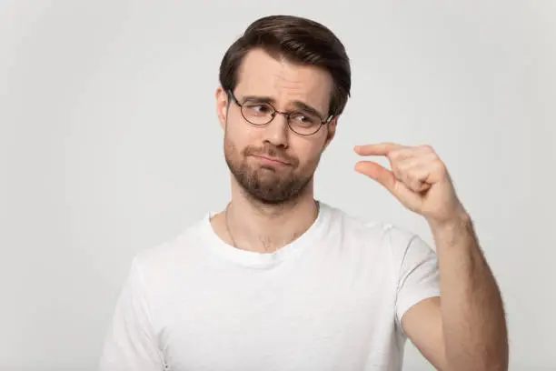 Head shot portrait guy isolated on grey background wearing glasses look at hand showing with fingers something small feels disappointed pity about little size insufficient length or thickness concept.
