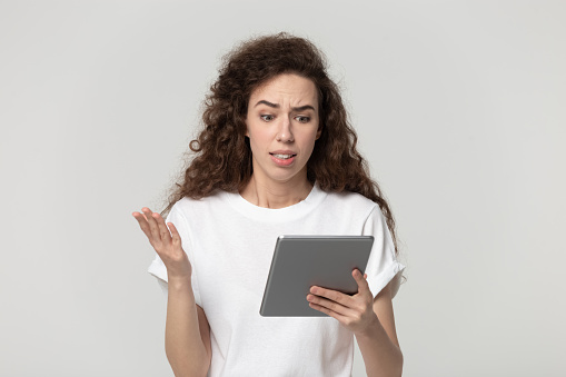 Irritated young woman looking at broken tablet screen gesticulates feels anger and annoyance having problems with gadget or received unpleasant message bad news posing on grey background studio shot.
