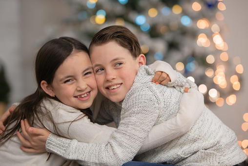 A caucasian sister and brother laugh and smile as they hug each other in the living room, in front of the Christmas tree.
