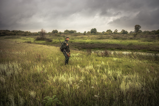 Hunting hunter men with gun standing in field nearby river during hunting season in overcast day with dramatic sky