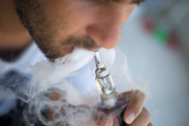 een man rookt een elektronische sigaret op een grijze achtergrond, blazen een stroom van rook - vape stockfoto's en -beelden