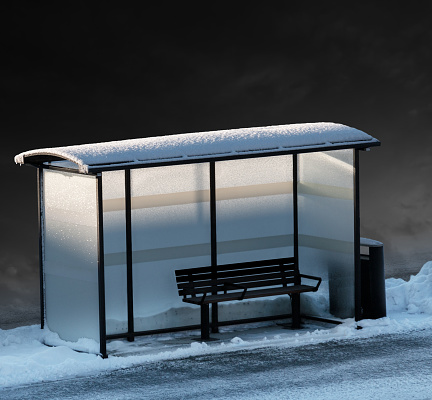 Empty bench in bus stop in winter, with dark sky in background and some dim light from low sun.
