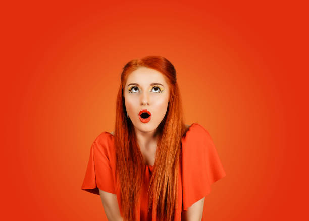 Portrait of young woman with shocked facial expression looking up to the copyspace isolated on red background