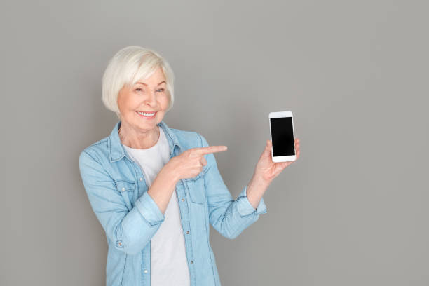 istock Senior woman in jeans jacket studio isolated on grey wall pointing at smartphone screen 1170598421