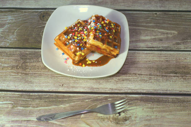 Two Viennese waffles with icing and topping, on a plate lie on a table near a fork.