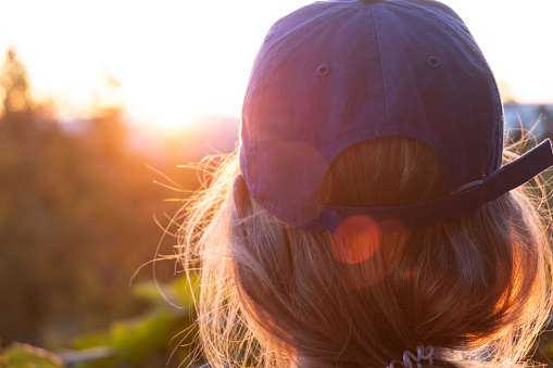A young Oregonian woman with blonde hair and a blue baseball cap looks out at the horizon as the sun peaks behind the mountains. The sun illuminates the edges of her hair and creates some beautiful lens flare.