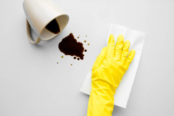 istock Hand in yellow protective rubber glove cleaning fresh spilled dark beverage. Coffee stain simple removing with white paper napkin. Cleanup. Close up. Gray background. Top view. 1170637231