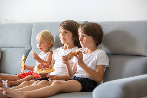 istock Cute children, sitting on couch, eating pizza and watching TV. Hungry child taking a bite from pizza on a pizza party 1170640468