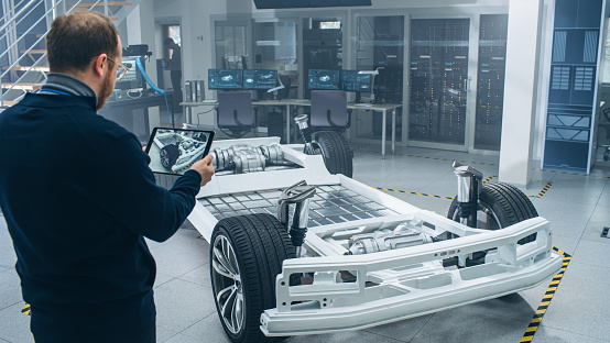 Engineer with Glasses and Beard Scans Electric Car Chassis Prototype with Wheels, Batteries and Engine with an Augmented Reality Software on a Tablet Computer in a High Tech Development Laboratory.