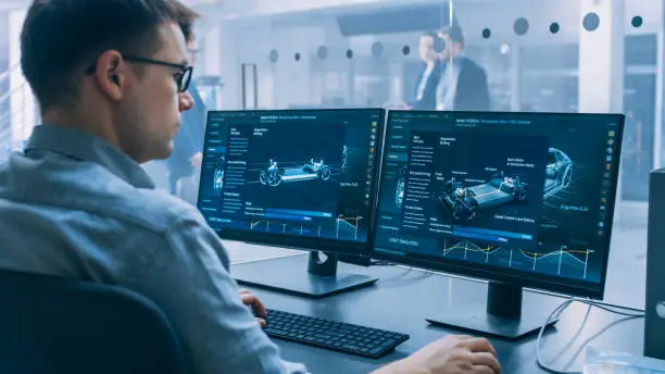 Professional Engineer Works on a Computer with a 3D CAD Software and Tests the Electric Car Chassis Prototype with Wheels, Batteries and Engine Standing in a High Tech Development Laboratory.