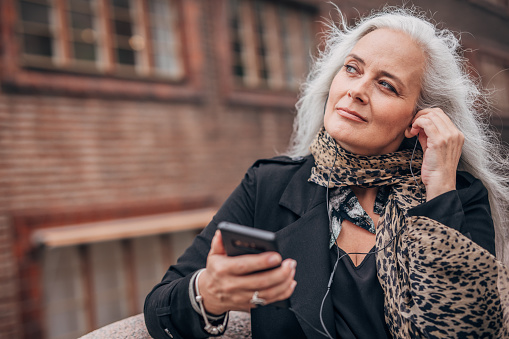 One woman, modern mature lady with gray hair, listening music on headphones, using smart phone.