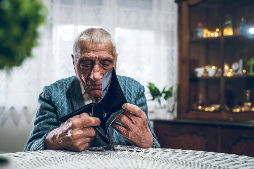 Old man looking at his empty wallet, sitting at the table, home interior. Day.