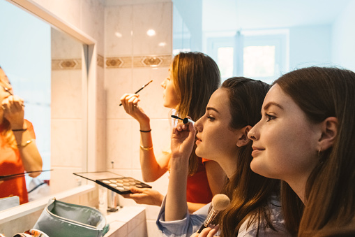 three young woman putting up make up in front of mirror in bath room