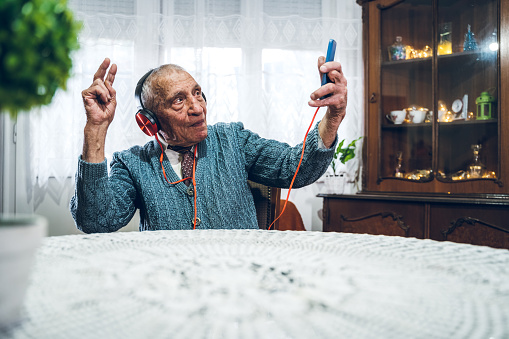 Senior, happy, man listening to the music, with headphones on. Using smart phone. Making happy gestures. Home interior.