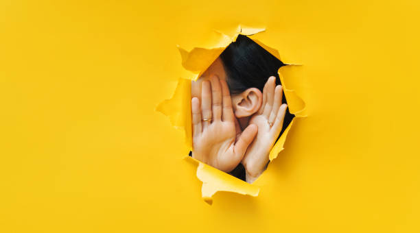 Female ear and hands close-up. Copy space. Torn paper, yellow background. The concept of eavesdropping, espionage, gossip and the yellow press. stock photo